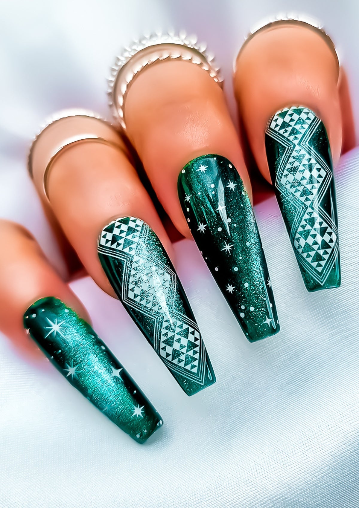 Long coffin shaped nails in emerald cat eye gel polish with white Maori nail art in Aonui design by Adrienne Whitewood on the index and ring fingers, and stars on the pinky and middle fingers. 